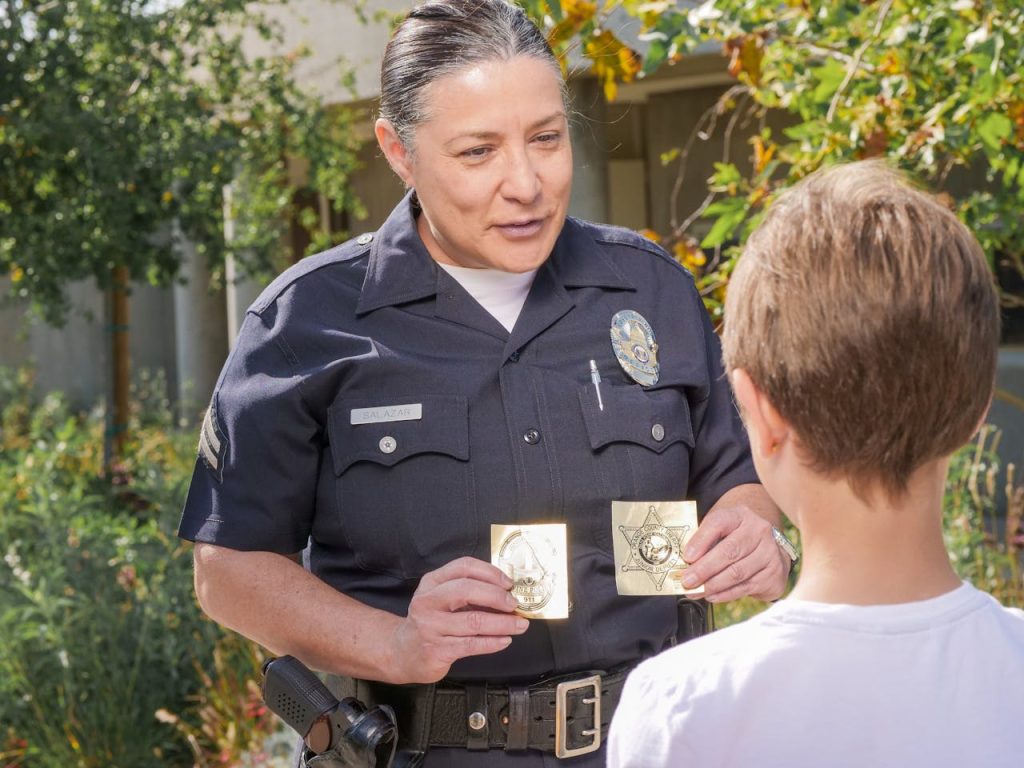 officer-showing-police-badges-to-a-young-boy-7715075 Friendly police officer showing badges to a child outdoors in a community setting.